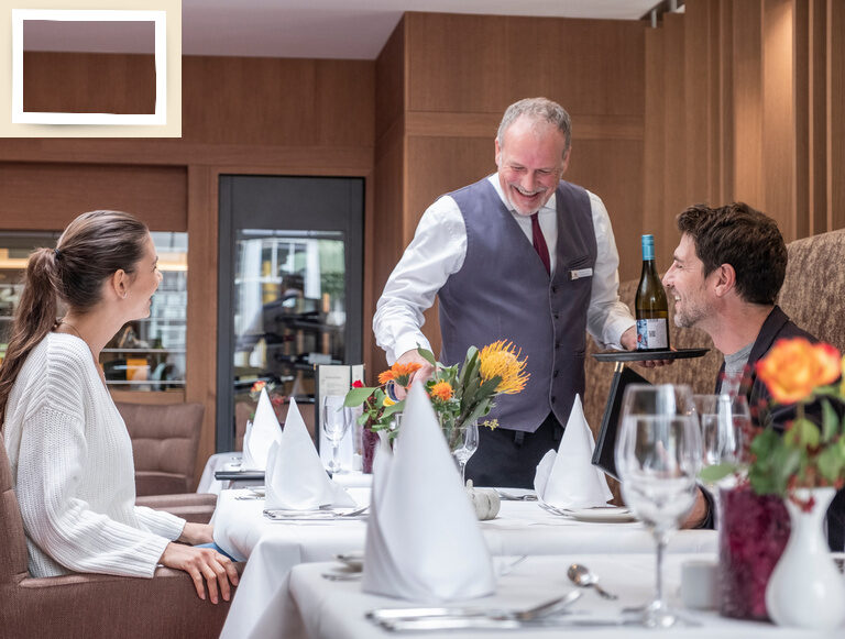 Zwei Personen genießen ein Dinner in der Weinlounge des Hotel Holzapfel im Rottal mit erlesenen ausgeschenkten Weinen von einem Kellner an einem fein gedeckten Tisch.